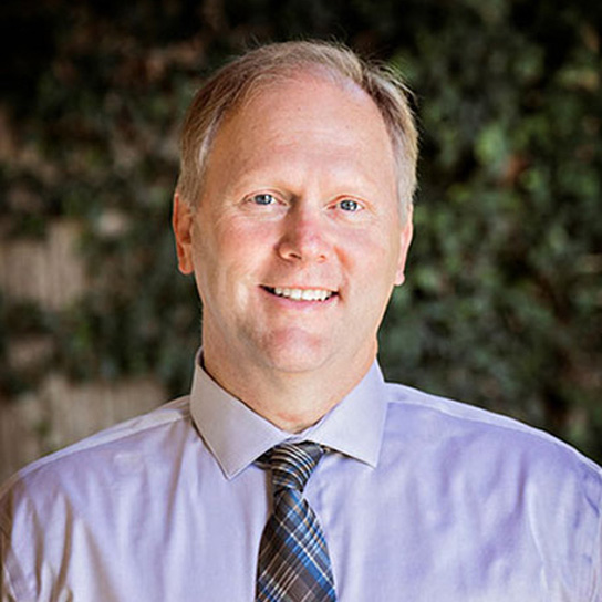 Brent Bell, DDS, smiling dentist in professional attire, with greenery in the background, representing Linrose Dental in Saratoga Springs.