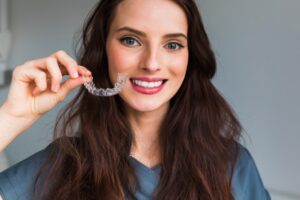 Smiling woman holding Invisalign clear aligners, showcasing the benefits of clear aligners in dental consultations at Linrose Dental in Saratoga Springs, UT.