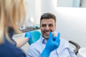 Man in dental chair smiling while holding clear aligner, with dental professional in blue gloves demonstrating Invisalign treatment process at Linrose Dental.